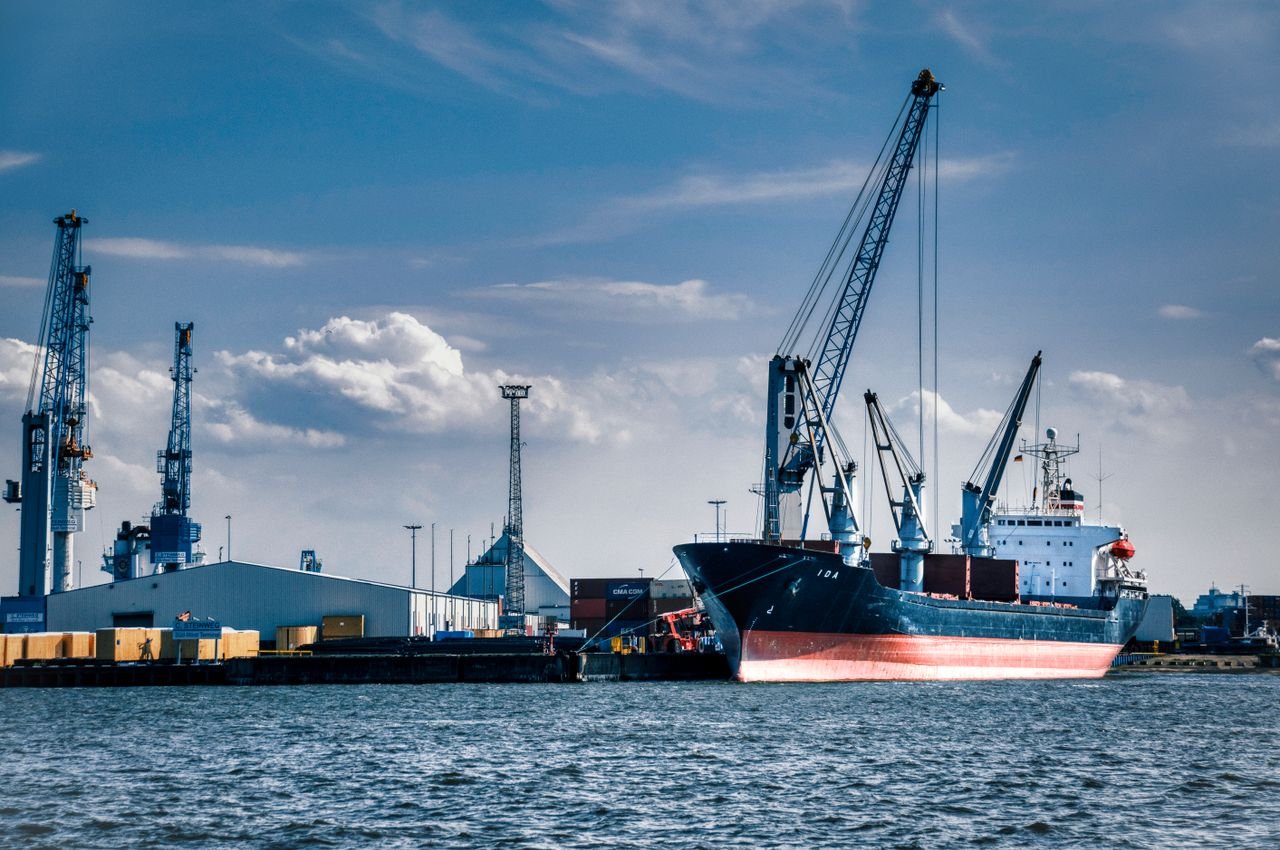 A vibrant scene of a cargo ship loaded with containers at a busy harbor under a clear blue sky.